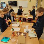 Dzieci w klasie przeprowadzają eksperyment naukowy, używając wagi, lejków i kubków pomiarowych przy swoich biurkach.