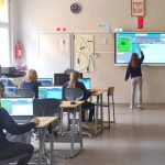 Uczniowie siedzą przy biurkach z laptopami, podczas gdy dziewczyna pisze na dużej interaktywnej tablicy w klasie.