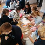 Grupa dzieci siedzi na podłodze, pracując razem nad rysunkami przy użyciu kolorowych markerów i papieru.