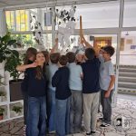 Grupa dzieci w szkolnych mundurkach gromadzi się wokół białego drzewa z karteczkami przyczepionymi do gałęzi.