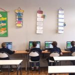 Pięciu uczniów siedzi przy biurkach, korzystając z komputerów stacjonarnych w klasie z kolorowymi plakatami na ścianie.