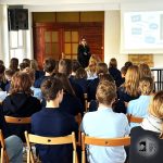 Kobieta prezentuje przed klasą siedzących uczniów, którzy stoją przed ekranem projektora.