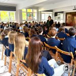 Uczniowie siedzący na drewnianych krzesłach stoją przed kobietą przemawiającą z przodu jasnej klasy.