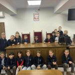 Grupa uczniów pozuje i uśmiecha się, siedząc na ławce sędziowskiej w sali sądowej.