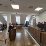 Młody chłopak stoi na podium w sali sądowej z siedzącymi ludźmi i sędziami na ławce.