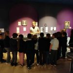 Grupa dzieci słucha przewodnika przed wystawą muzealną z fioletowymi ścianami.