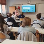 Uczniowie siedzą w ławkach naprzeciwko nauczyciela i ekranu wyświetlającego kolorowy slajd edukacyjny w klasie.