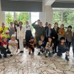 Grupa dzieci w kostiumach, pozujących razem w pomieszczeniu z dużymi oknami i drzewami widocznymi na zewnątrz.