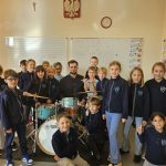 Grupa dzieci i dorosły stoją wokół zestawu perkusyjnego w klasie, uśmiechając się do kamery.