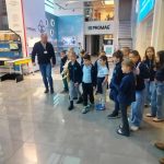 Grupa dzieci i dorosły przewodnik stoją wewnątrz nowoczesnego salonu wystawowego z wyświetlaczami i błyszczącymi podłogami.