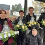 Pięcioro dzieci w kurtkach trzyma kompozycje kwiatowe z lampionami na przystanku autobusowym w deszczowy dzień.