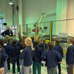 Grupa dzieci słucha mężczyzny objaśniającego maszyny w nowoczesnym zakładzie przemysłowym.