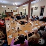 Grupa ludzi siedzących przy stołach, dzielących się jedzeniem i napojami w dobrze oświetlonym pomieszczeniu.