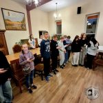 Grupa dzieci stoi w pokoju, niektóre trzymają telefony, ze stołem i krzesłami w tle.