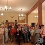 Grupa starszych i młodszych osób pozujących razem w ciepło oświetlonym pokoju z obrazami na ścianie.