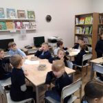 Dzieci siedzą przy stolikach w klasie z regałami, komputerami i książkami widocznymi w tle.