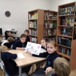 Dzieci w klasie siedzą przy stolikach, rysując i trzymając prace plastyczne, otoczone półkami z książkami i komputerem.