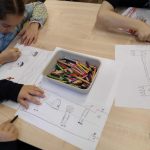 Troje dzieci rysuje kredkami na papierze przy stole; taca z kredkami znajduje się pośrodku.