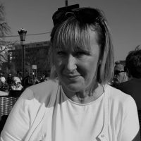 Kobieta w okularach przeciwsłonecznych siedzi na zewnątrz, uśmiechnięta, z ludźmi i budynkami w tle.