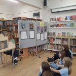 Kobieta czyta dzieciom siedzącym na podłodze w bibliotece, za którą znajdują się półki z książkami i tablice informacyjne.