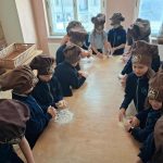 Dzieci w brązowych czapkach kucharskich gromadzą się wokół stołu, wyrabiając ciasto w dobrze oświetlonej klasie.