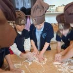 Dzieci w czapkach kucharskich ugniatają ciasto na posypanym mąką stole podczas zajęć kulinarnych.