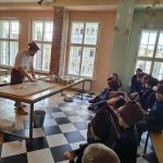 Szef kuchni demonstruje pieczenie grupie dzieci w brązowych kapeluszach w jasnym pokoju z podłogą w kratkę.