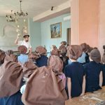 Grupa dzieci w brązowych kapeluszach słucha instruktora w klasie ozdobionej żyrandolami.