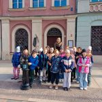 Grupa dzieci i dorosły pozują przed kolorowymi budynkami w słoneczny dzień.