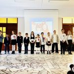 Dzieci stoją w rzędzie na scenie, prezentując przed ekranem w warunkach szkolnych.