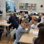 Uczniowie siedzą w grupach przy stolikach w bibliotece, niektórzy podnoszą ręce, angażując się w dyskusję lub ćwiczenia.