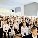 Grupa dzieci i nauczyciel siedzą w pomieszczeniu, ubrani formalnie, uczestnicząc w szkolnym wydarzeniu lub zebraniu.