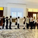 Grupa dzieci w mundurkach stoi w kolejce przed ekranem z tekstem i obrazami w otoczeniu szkolnym.