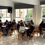 Uczniowie siedzą w grupach przy stołach w jasnej sali lekcyjnej z dużymi oknami, pracując wspólnie nad zadaniami.