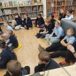 Grupa dzieci siedzi na podłodze i kanapie w bibliotece, uważnie słuchając.