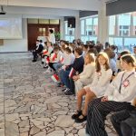 Uczniowie siedzą w rzędach, niektórzy trzymają polskie flagi, oglądając prezentację na ekranie z przodu.