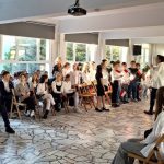 Dzieci w oficjalnych strojach siedzą i stoją w jasnym pomieszczeniu, uczestnicząc w szkolnym wydarzeniu lub prezentacji.