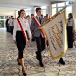 Trzech uczniów w oficjalnych strojach niesie sztandar szkoły podczas ceremonii, a inni stoją w szeregu.