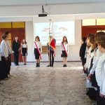 Uczniowie w oficjalnych strojach stoją w dwóch rzędach naprzeciwko siebie podczas szkolnego apelu z flagami pośrodku.