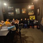 Grupa dzieci siedzi w ciemnym pokoju, słuchając prezentera trzymającego dużą tablicę z tekstem.