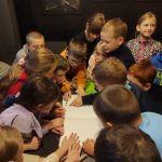 Grupa dzieci gromadzi się wokół stołu, patrząc razem na otwartą książkę w słabo oświetlonym pokoju.