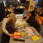 Siedmioro małych dzieci gromadzi się wokół stołu, pracując razem nad kolorowym projektem artystycznym i rzemieślniczym.