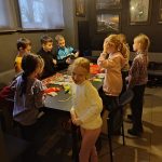 Ośmioro dzieci pracuje razem przy stole w przytulnym, słabo oświetlonym pokoju z obrazami na ścianach.