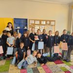 Grupa dzieci i nauczyciel uśmiechają się, trzymając torby z prezentami w kolorowej klasie z rysunkami na ścianie.