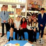 Sześcioro nastolatków i jeden dorosły pozują razem w pomieszczeniu, uśmiechnięci, z tablicami i zdjęciami za sobą.