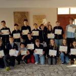 Grupa uczniów i nauczycieli w pomieszczeniu, trzymająca certyfikaty i pozująca do zdjęcia grupowego.