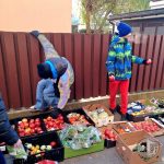 Dwóch chłopców w zimowych ubraniach przy płocie; jeden wspina się, podczas gdy drugi trzyma jabłka w pobliżu skrzynek z produktami.