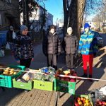 Czworo dzieci w zimowych ubraniach stoi za stołem z owocami i warzywami na targu w słoneczny dzień.