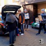 Ludzie rozładowują żywność i zapasy z samochodu; zakonnica i trzech chłopców pomaga w dystrybucji.