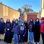 Grupa ludzi, w tym zakonnica i dzieci w zimowych ubraniach, stoi na zewnątrz przy beżowym budynku.